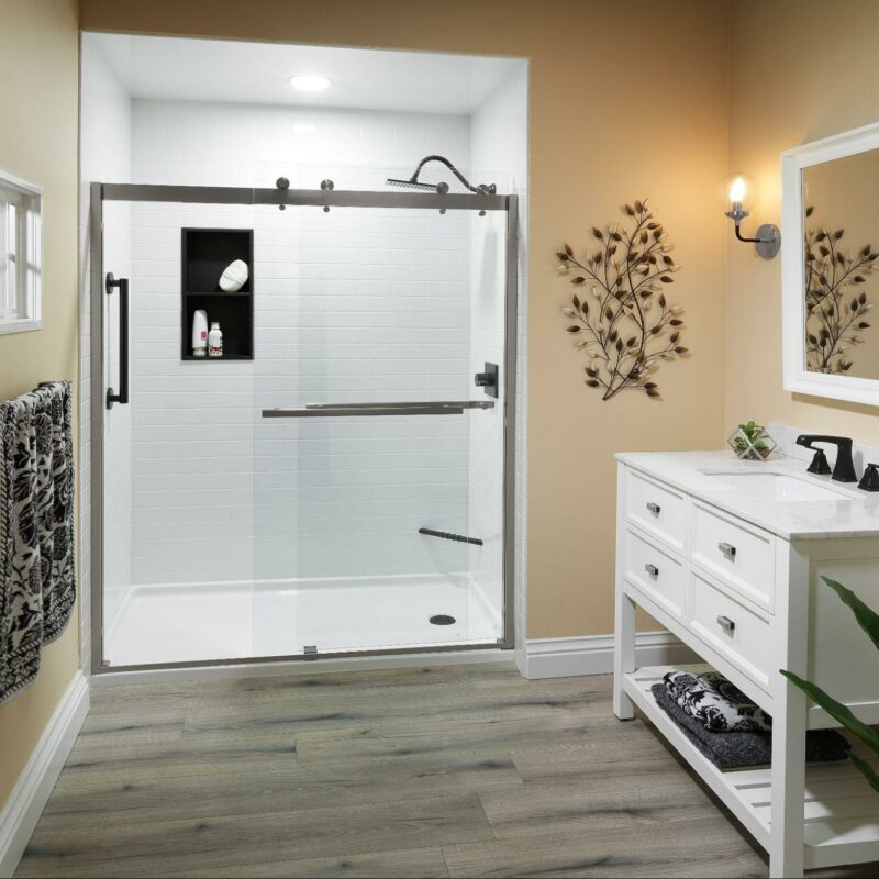 A modern bathroom featuring a walk-in shower with a glass door, white subway tiles, and a recessed shelf. The vanity has a white marble countertop, double sinks with black faucets, and a matching white wooden frame around the mirror. Decor includes two wall-mounted leafy metal artworks, a potted plant, and black and white patterned towels. The floor is covered with grey wood-like tiles.